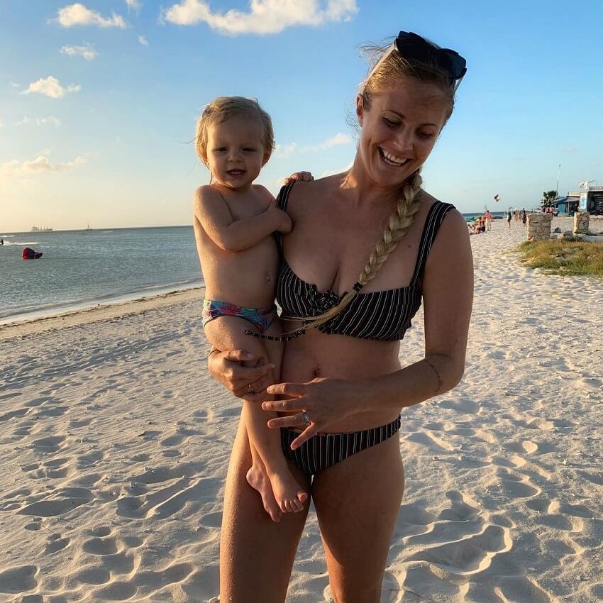 Rachel Brathen smiling on the beach while holding her baby in her arms, looking fit and lean